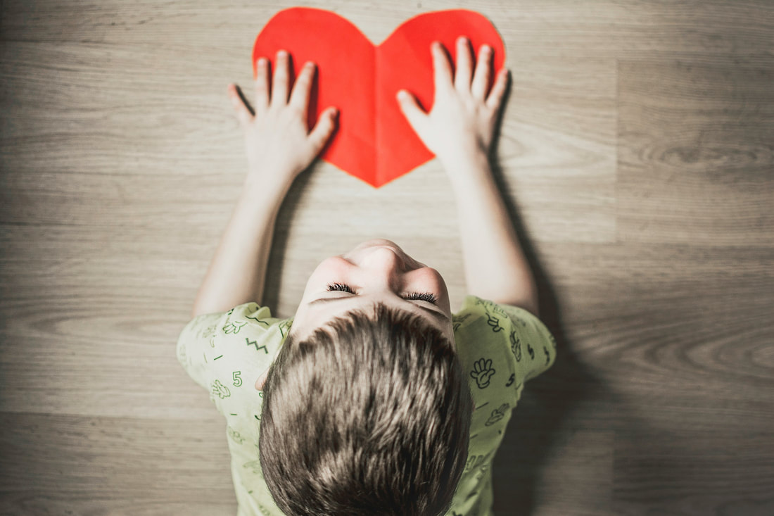 Blonde male child laying down on a brown hardwood floor holding a red cut-out paper heart