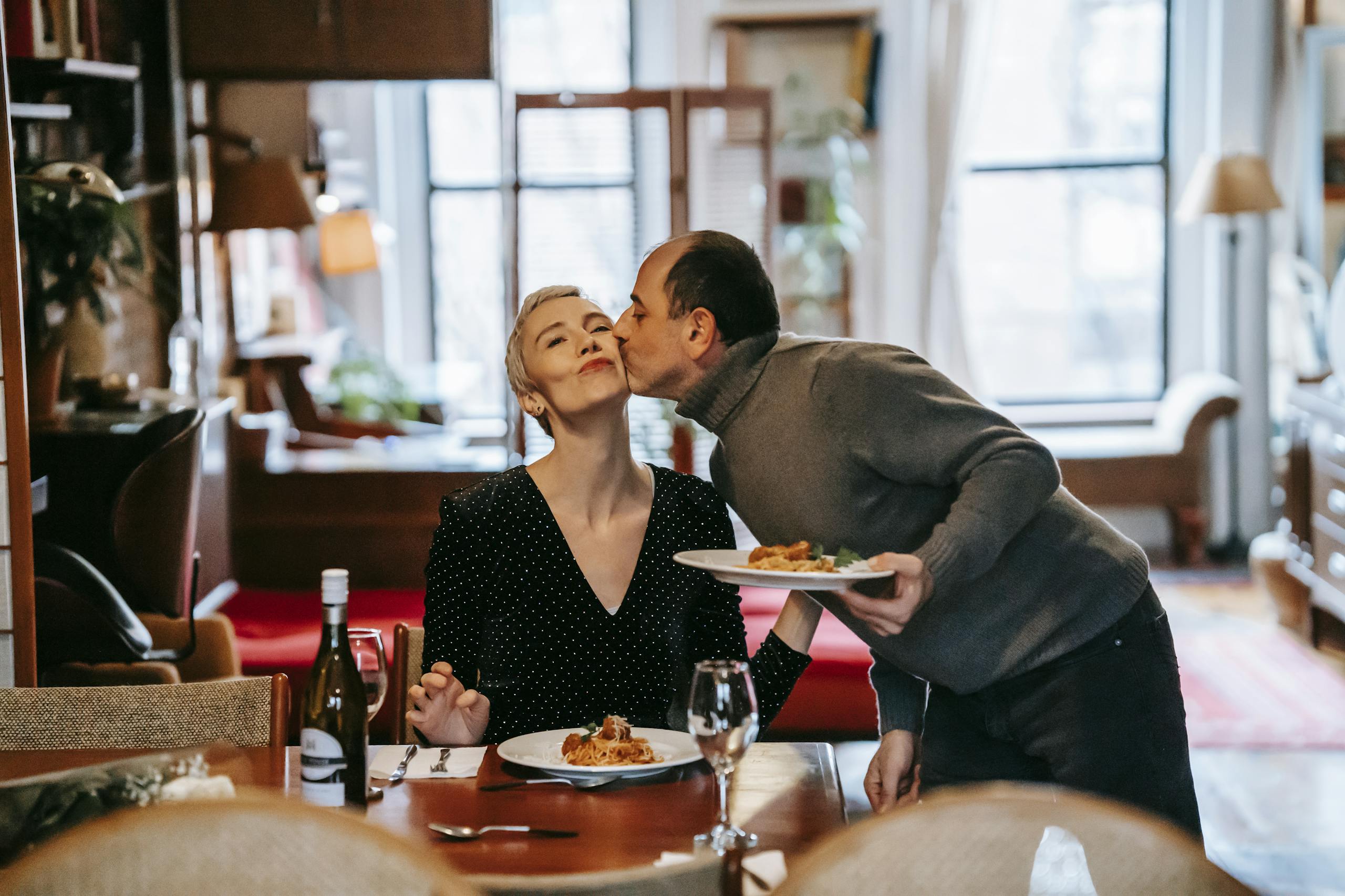 Gentle husband with plate of meal kissing wife having romantic dinner with bottle of wine at home